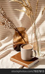 still life, hygge and drinks concept - cup of coffee with cinnamon on book, decorative dried flowers in vase and bottle with drapery. cup of coffee on book and dried flowers in vases