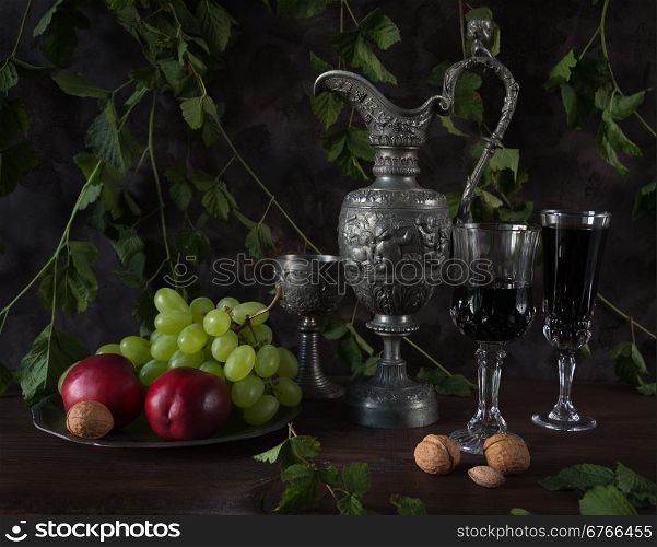 Still life in medieval style with antique pitcher and a goblets filled with red wine as well as walnuts, green grapes and nectarines on a dark background, covered with green plants