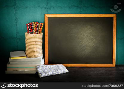 Still life of empty blackboard and colorful pencils with books on wooden table over green wall background.