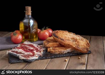 Still life of Iberian ham with tomato bread and extra olive oil on slate stone tray. Black background.