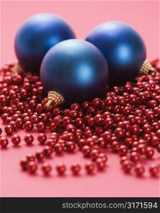 Still life of large blue Christmas ornaments and strings of red beads on red background.