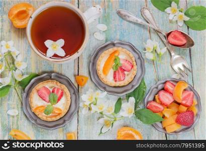 Still-life with a cup of black tea, tartlets filled with cream and berries and jasmine flowers on old shabby boards, top view