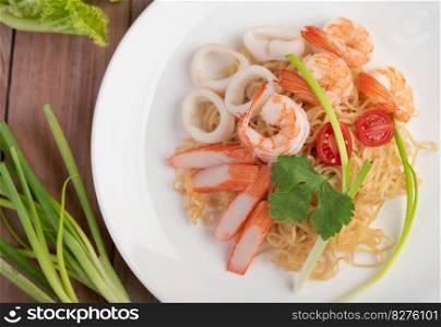 Stir-fried instant noodle with prawn and crab stick along with tomatoes and peppers in a white plate on a wooden floor.
