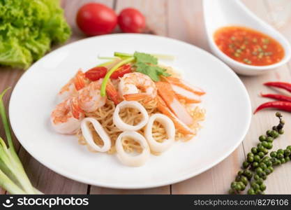 Stir-fried instant noodle with prawn and crab stick along with tomatoes and peppers in a white plate on a wooden floor.