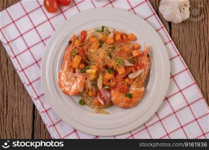 Stir-fried prawns with glass noodles in a white plate placed on a cloth with eggs and garlic.