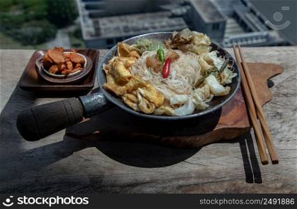 Stir fried rice vermicelli with pork dumplings, egg and vegetable in small steaming iron pot serve with crispy pork crackling or pork scratching on rustic old wooden table. Asia food style, Selective focus.