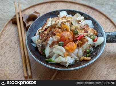 Stir-fried wide rice noodles with Bacon, Egg, Dried shrimp in small steaming iron pot and wooden chopsticks served on round wooden tray. Selective focus.