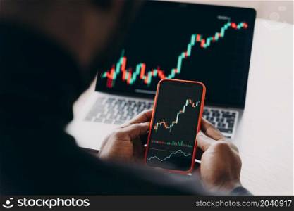 Stock trading concept. Cropped photo of confident Afro American businessman holding smartphone, works with financial statistics, comparing charts diagrams on phone and laptop screens, analyzing data. Confident Afro American stock trader holding smartphone, works with financial statistics online