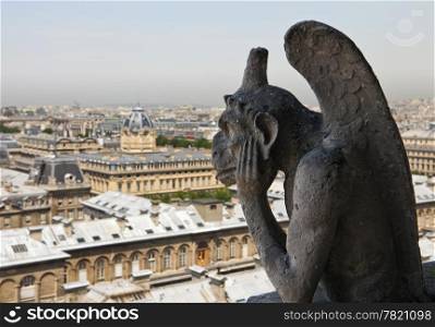Stone carving of a French gargoyle is looking over the city of Paris while thinking and holding its head in its hands.