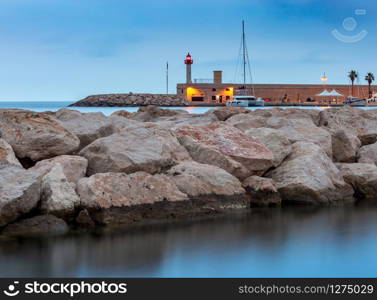 Stone lighthouse at the breakwater at the entrance to the port in night lighting. Menton. France.. Menton. Stone lighthouse on the breakwater at the entrance to the port.