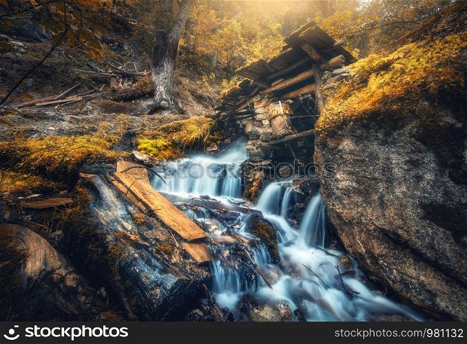 Stony well in colorful forest with little waterfall in mountain river at sunset in autumn. Landscape with stones in water, building, orange trees, waterfall and vibrant foliage in fall. Nature. Stony well in colorful orange forest with little waterfall