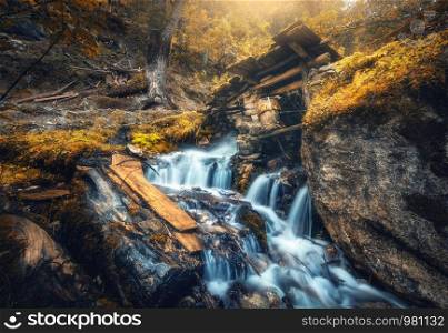 Stony well in colorful forest with little waterfall in mountain river at sunset in autumn. Landscape with stones in water, building, orange trees, waterfall and vibrant foliage in fall. Nature. Stony well in colorful orange forest with little waterfall
