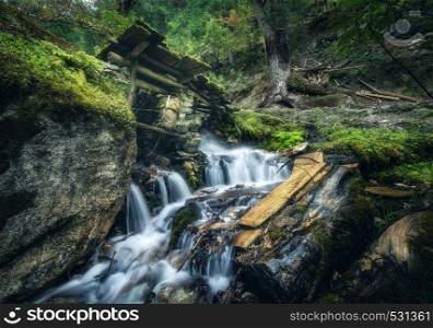 Stony well in colorful green forest with little waterfall in mountain river at sunset in summer. Landscape with stones in water, building, trees, waterfall and vibrant foliage. Nature. Blurred water