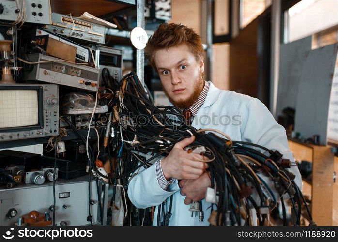 Strange engineer holds bunch of wires with different plugs in laboratory. Electrical testing tools on background. Lab equipment, engineering workshop