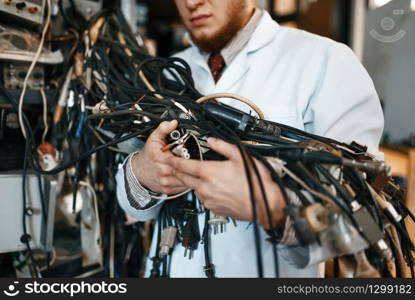 Strange engineer holds bunch of wires with different plugs in laboratory. Electrical testing tools on background. Lab equipment, engineering workshop. Engineer holds bunch of wires in laboratory