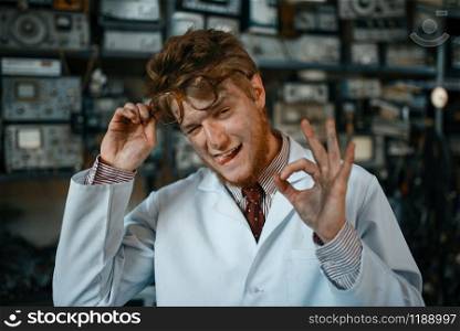 Strange male scientist shows OK sign in laboratory. Electrical testing tools on background. Lab equipment, engineering workshop