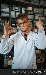 Strange male scientist shows signs with his fingers in laboratory. Electrical testing tools on background. Lab equipment, engineering workshop