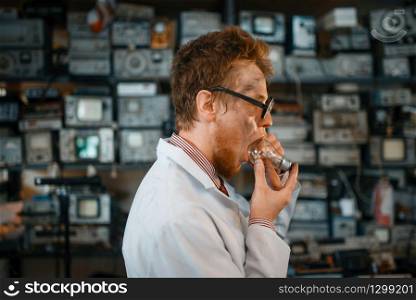 Strange scientist trying to put a light bulb in his mouth, test in laboratory. Electrical testing tools on background. Lab equipment, engineering workshop