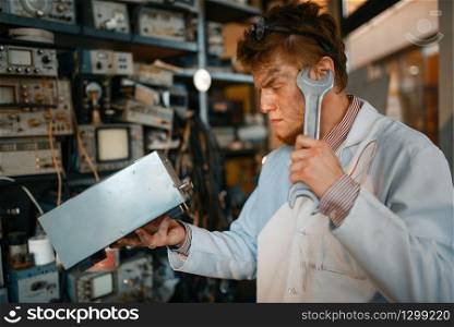 Strange scientist with electric device and huge wrench, test in laboratory. Electrical testing tools on background. Lab equipment, engineering workshop