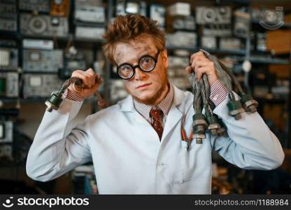 Strange scientist with electric terminal connected to his ear, dangerous test in laboratory. Electrical testing tools on background. Lab equipment, engineering workshop. Scientist with terminal connected to his ear, test