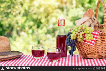 Straw hat, basket with grapes and juice on a red checkered tablecloth. Natural green background. Picnic concept. Straw hat, basket with grapes and juice on red checkered tablecloth