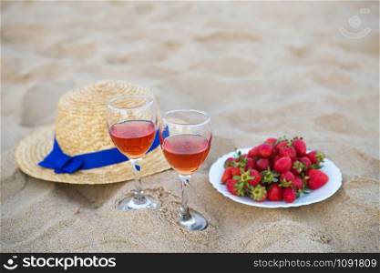 straw hat, two glasses of wine and a plate of strawberries on a sandy beach, close-up.. straw hat, two glasses of wine and a plate of strawberries on a sandy beach, close-up