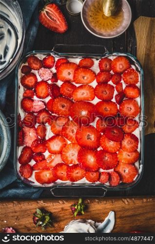 Strawberries cake preparation in glass dish on rustic kitchen table background, top view