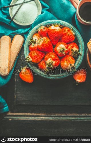 Strawberries for tiramisu making. Ripe strawberries in blue bowl with tiramisu cooking ingredients on old dark wooden background, top view, place for text. Italian food concept