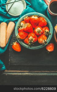 Strawberries for tiramisu making. Ripe strawberries in blue bowl with tiramisu cooking ingredients on old dark wooden background, top view, place for text. Italian food concept