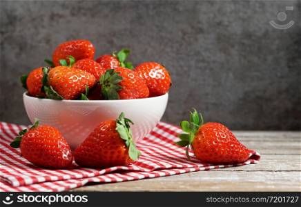 Strawberry concept with a group of ripe red strawberries in a white bowl close up frontal view on a vintage rustic wooden table and dark grey background.