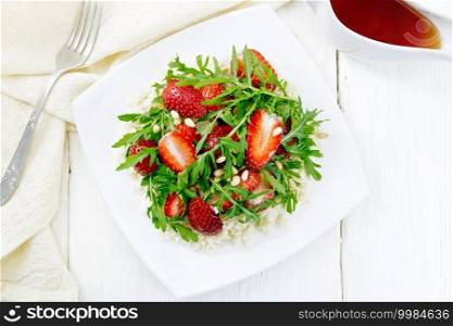 Strawberry, couscous, cedar nuts and arugula salad dressed with balsamic vinegar and olive oil in a plate, napkin and fork on wooden board background from above