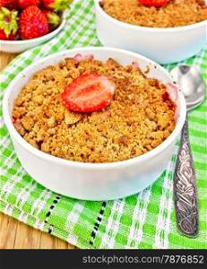 Strawberry crumble in a white bowl with a spoon on a green napkin, strawberries on a wooden boards background