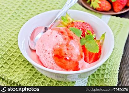 Strawberry ice cream in a white bowl with strawberries, strawberry syrup and a spoon on a green napkin on a wooden boards background