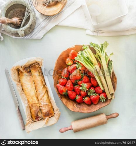 Strawberry rhubarb wrap cake on kitchen table background with bowls with ingredients. Top view. Summer baking with seasonal products . Home cuisine