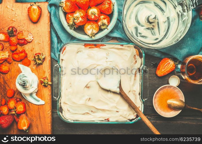 Strawberry tiramisu cake making with cake dish , wooden spoon and mascarpone cream on dark rustic background, top view. Italian food concept