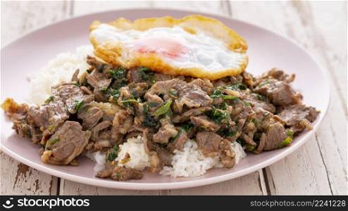 streamed rice with stir fried beef with tree basil leaves, long pepper, Indian long pepper, Javanese long pepper, pepper, garlic, chili and fried egg in ceramic plate on white wood texture background