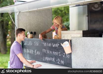 street sale and people concept - happy young saleswoman at food truck serving male customer and pointing finger at menu on chalkboard. saleswoman at food truck serving male customer