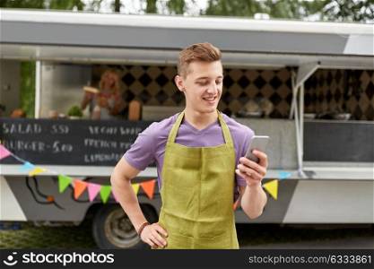 street sale, technology and people concept - happy young salesman in apron with smartphone at food truck. salesman in apron with smartphone at food truck