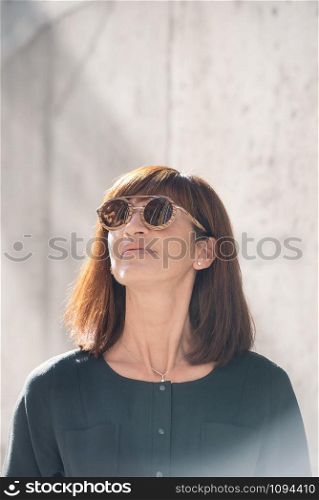 Street style Fashion portrait pretty middle age woman in stylish dark overall garment, smiling, sun glasses with wooden frame. Sunny day, city lifestyle