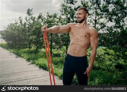 Strength and motivation concept. Muscular man with naked torso, has exercises with resistance band, performs training for biceps, looks into distance, poses outdoor. Sportsman uses expander.