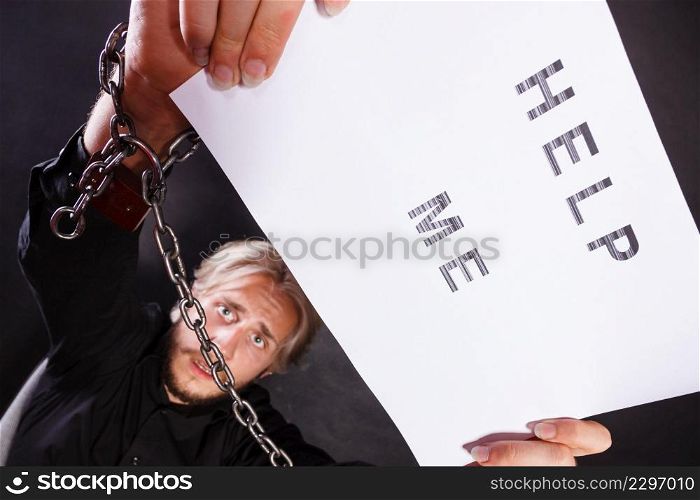 Stress, depression, assistance concept. Scared man with chained hands holding help me sign, studio shot on dark, grunge background. Man with chained hands holding help me sign