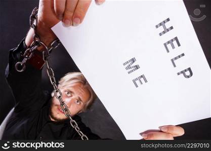 Stress, depression, assistance concept. Scared man with chained hands holding help me sign, studio shot on dark, grunge background. Man with chained hands holding help me sign