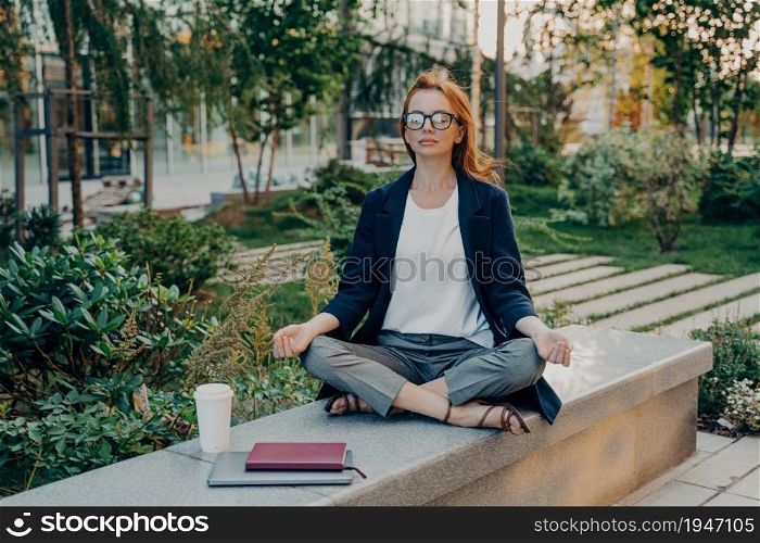 Stress free break and long working hours. Relaxed redhead young woman does yoga outside sits in lotus pose poses in park with takeaway coffee notepad and digital device near breathes deeply.. Relaxed redhead young woman does yoga outside sits in lotus pose poses in park