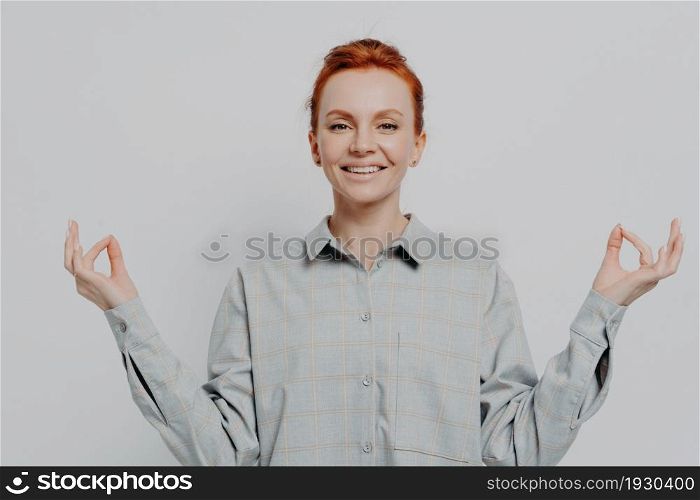 Stress free. Peaceful young smiling redhead woman in casual outfit isolated on grey studio background holding hands in mudra gesture and meditating, feeling calm while practicing yoga. Peaceful young smiling redhead woman holding hands in mudra isolated on grey studio background