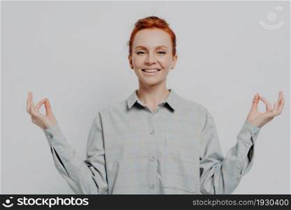 Stress free. Peaceful young smiling redhead woman in casual outfit isolated on grey studio background holding hands in mudra gesture and meditating, feeling calm while practicing yoga. Peaceful young smiling redhead woman holding hands in mudra isolated on grey studio background