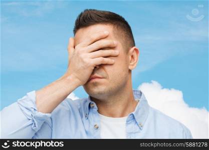 stress, headache, health care and people concept - unhappy man covering his eyes by hand over blue sky and cloud background