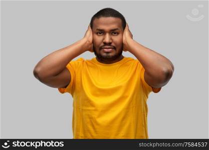 stress, noise and people concept - stressed young african american man in yellow t-shirt closing his ears with hands over grey background. stressed man closing his ears with hands