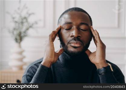 Stressed African home office worker sits at desk touching temples while eyes closed, feels tired out from hours of work online, suffers from migraine, bushed employee working on difficult task concept. Stressed African home office worker sits at desk touching temples while eyes closed