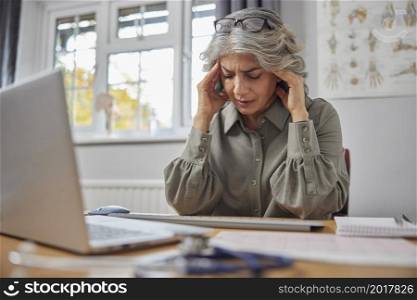 Stressed And Overworked Mature Female GP At Desk In Doctors Office