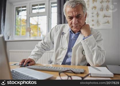 Stressed And Overworked Mature Male GP Wearing White Coat At Desk In Doctors Office